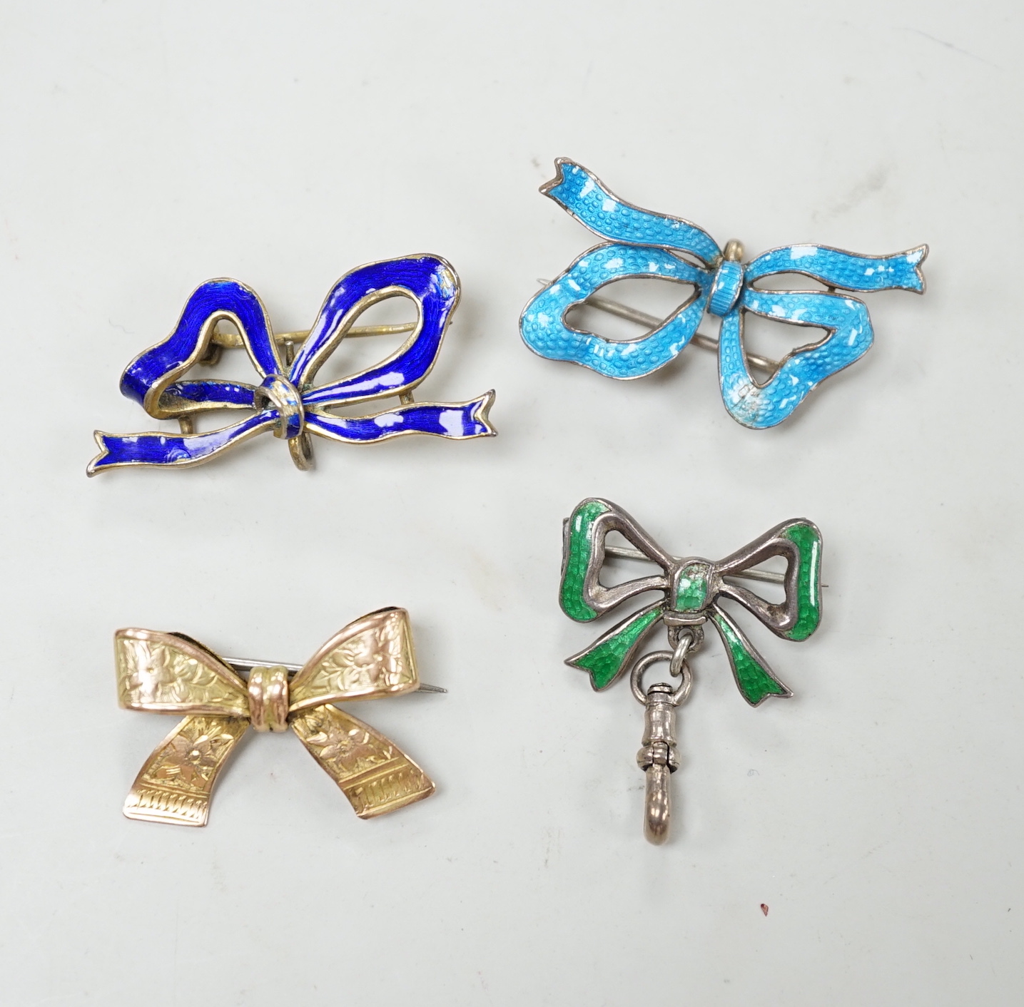 An engraved yellow metal ribbon bow brooch, 26mm, 2.8 grams, together with three silver/gilt white metal and enamel ribbon bow brooches.